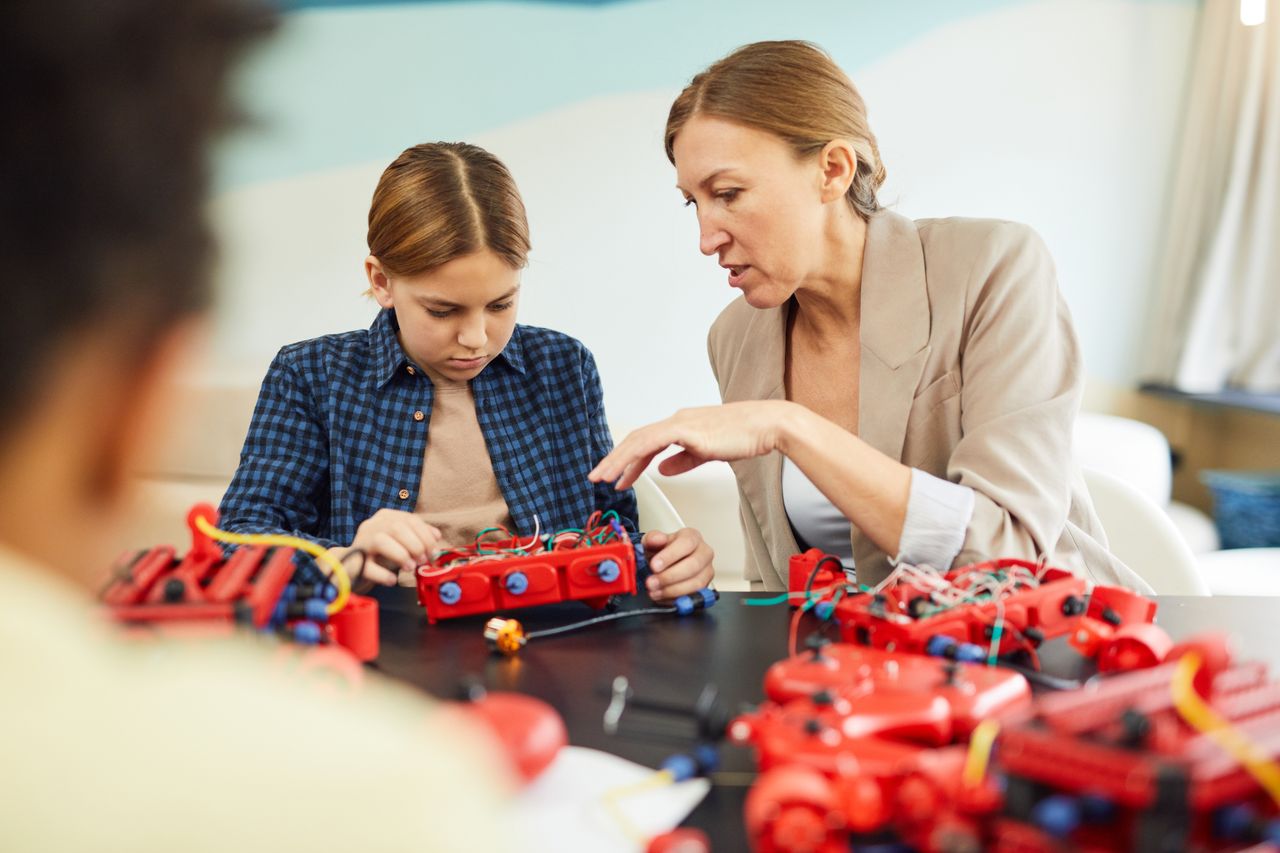 Services A woman educator helps a young student with a robotics project in a technology classroom.